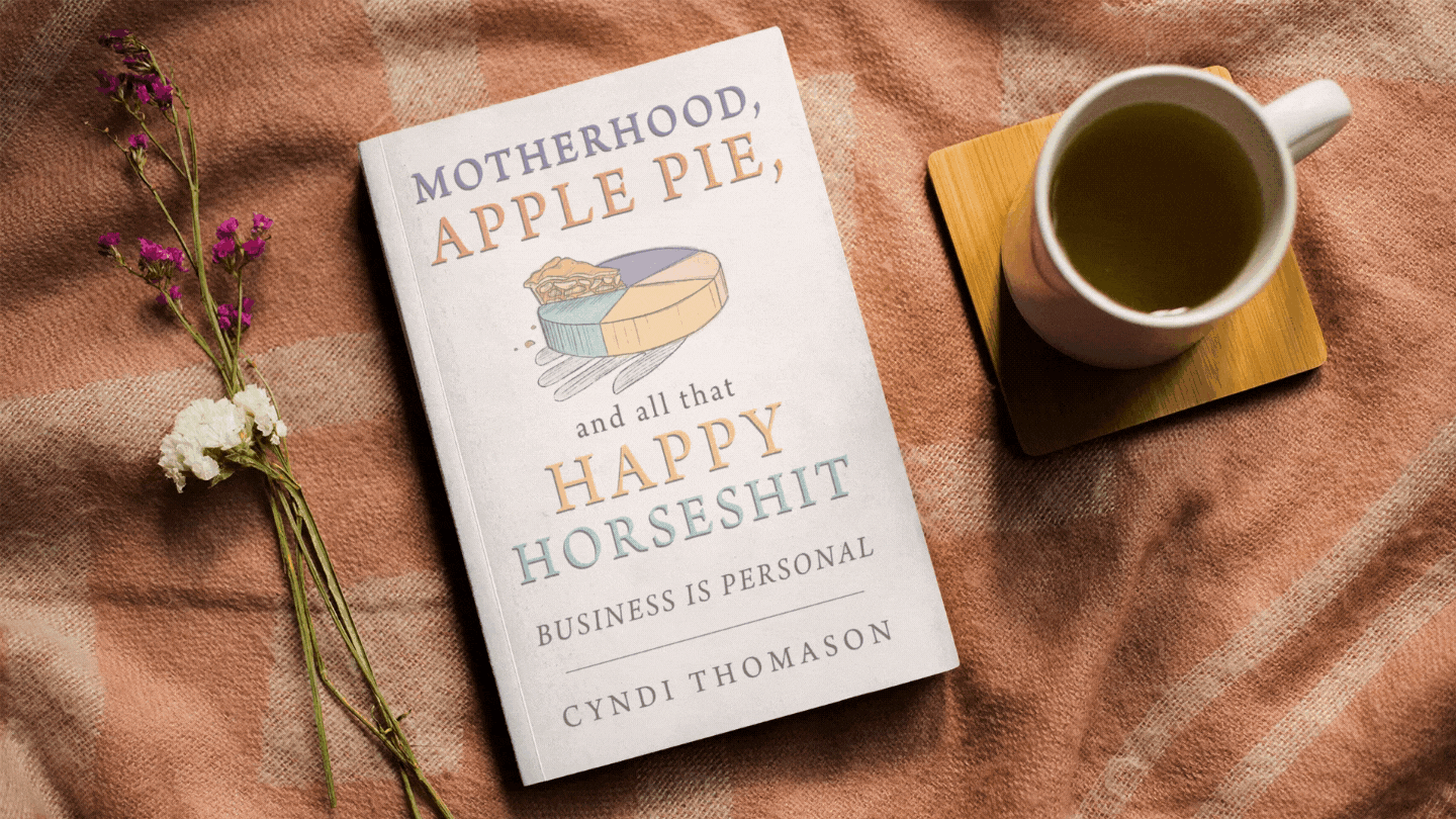 Image with the book Motherhood, Apple Pie, And All That Happy Horseshit on a table with a yellow mug of tea. 